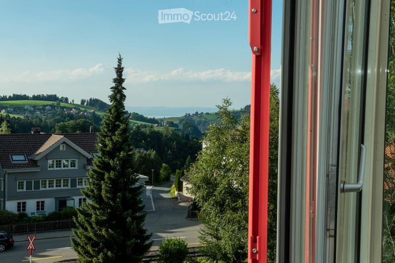 View from a balcony showing a suburban neighborhood with houses, trees, and a distant landscape under a clear sky.