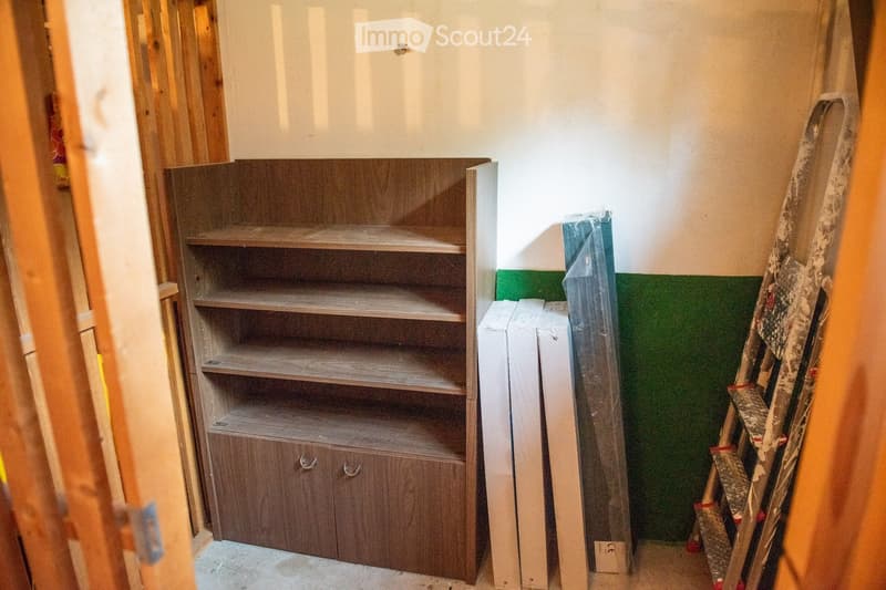 Empty room with unfinished walls, a wooden shelf unit, and construction materials including a ladder and plastic-wrapped items.
