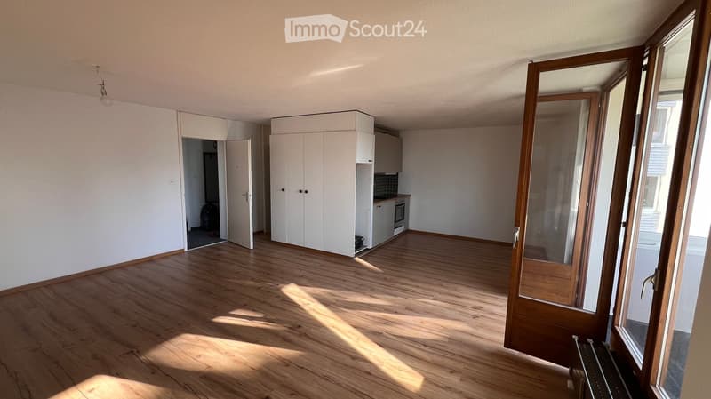 Empty room with wooden flooring, white walls, and large windows. The room is connected to a small kitchen area with white cabinets and an oven.