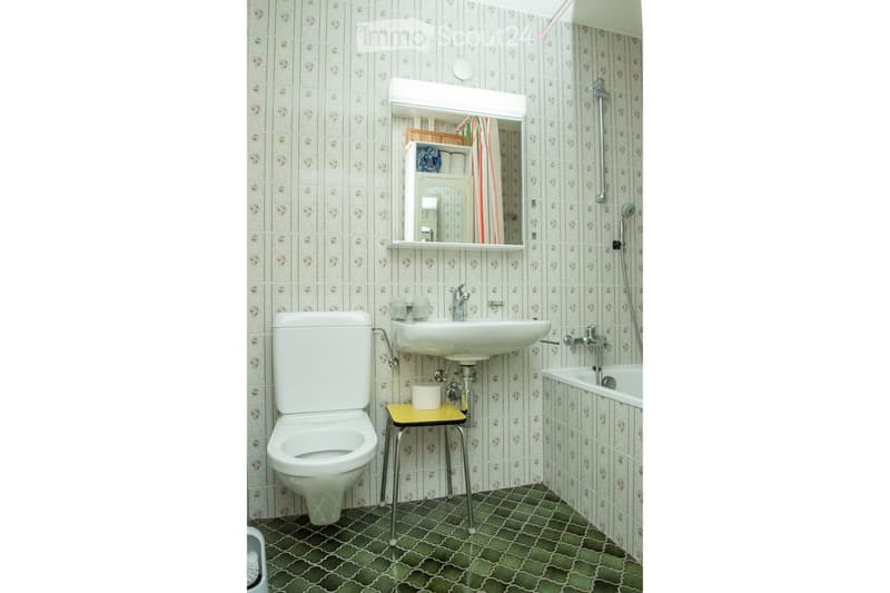 Bathroom with a toilet, sink, bathtub, mirror, and a small yellow stool. The walls are tiled with a light green pattern, and the floor has a green and white geometric tile design.