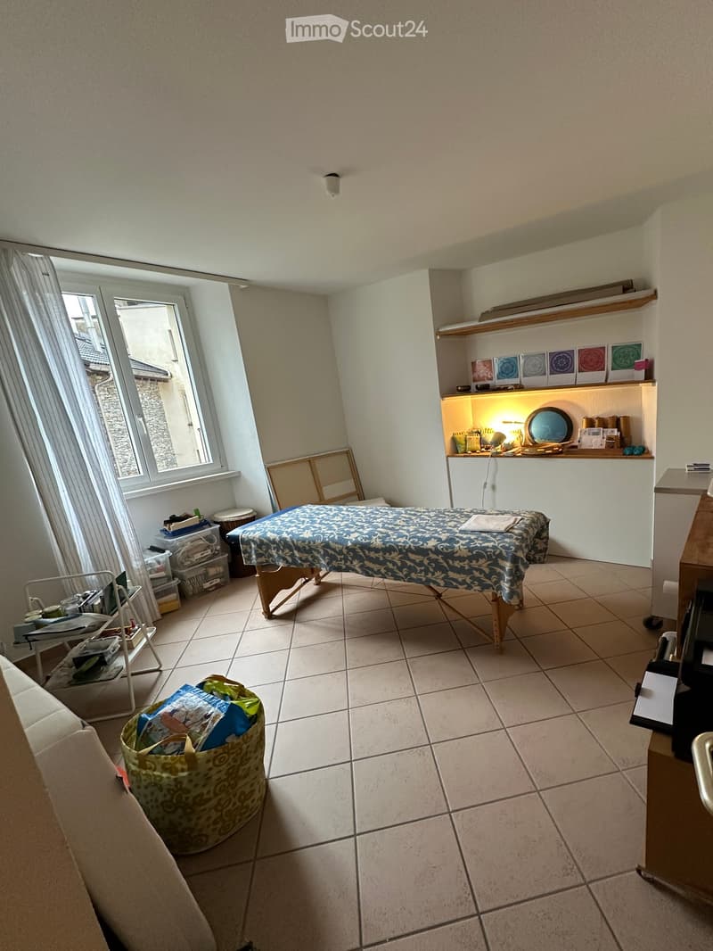 A furnished room with a massage table, shelves, a sofa, and storage bins. The room has tiled flooring and large windows with curtains.