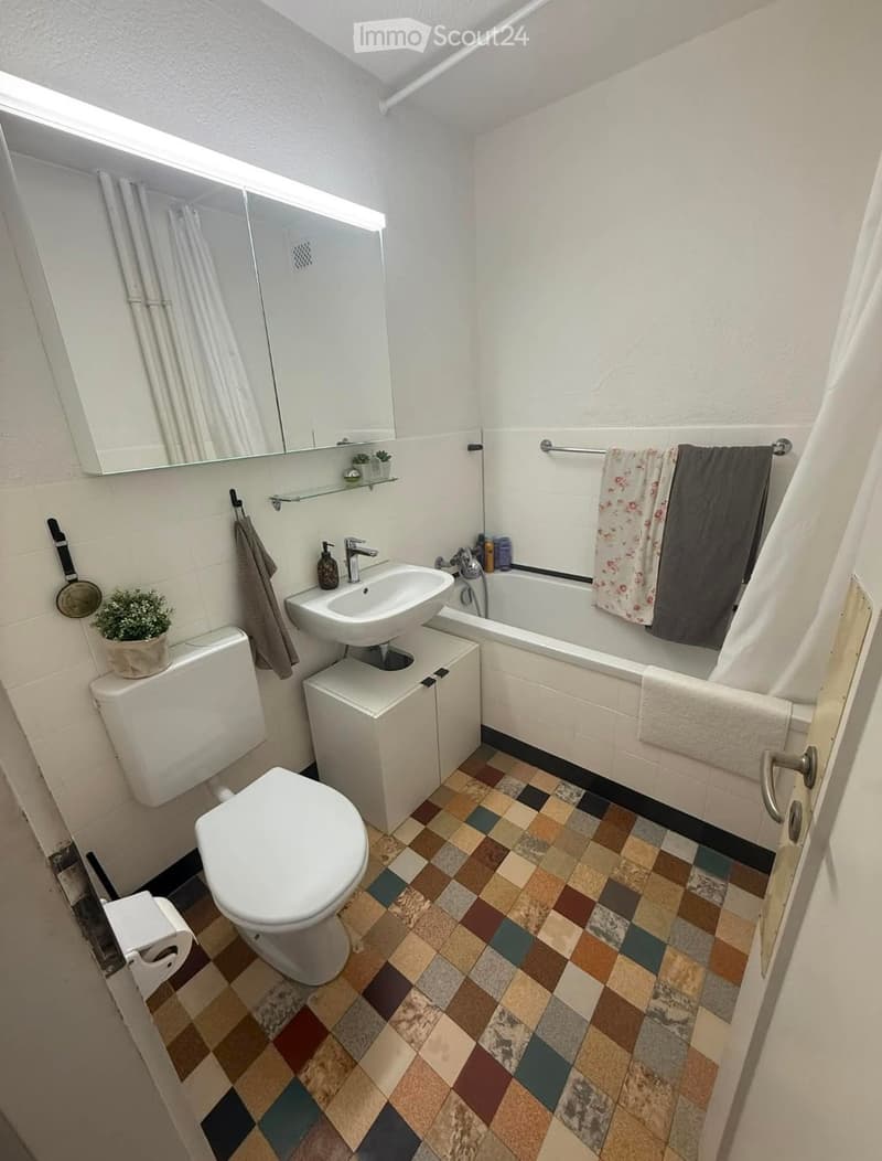 Modern bathroom with a white toilet, sink, bathtub, and mirror. The floor features a colorful mosaic tile design, and towels are neatly hung on a rack.