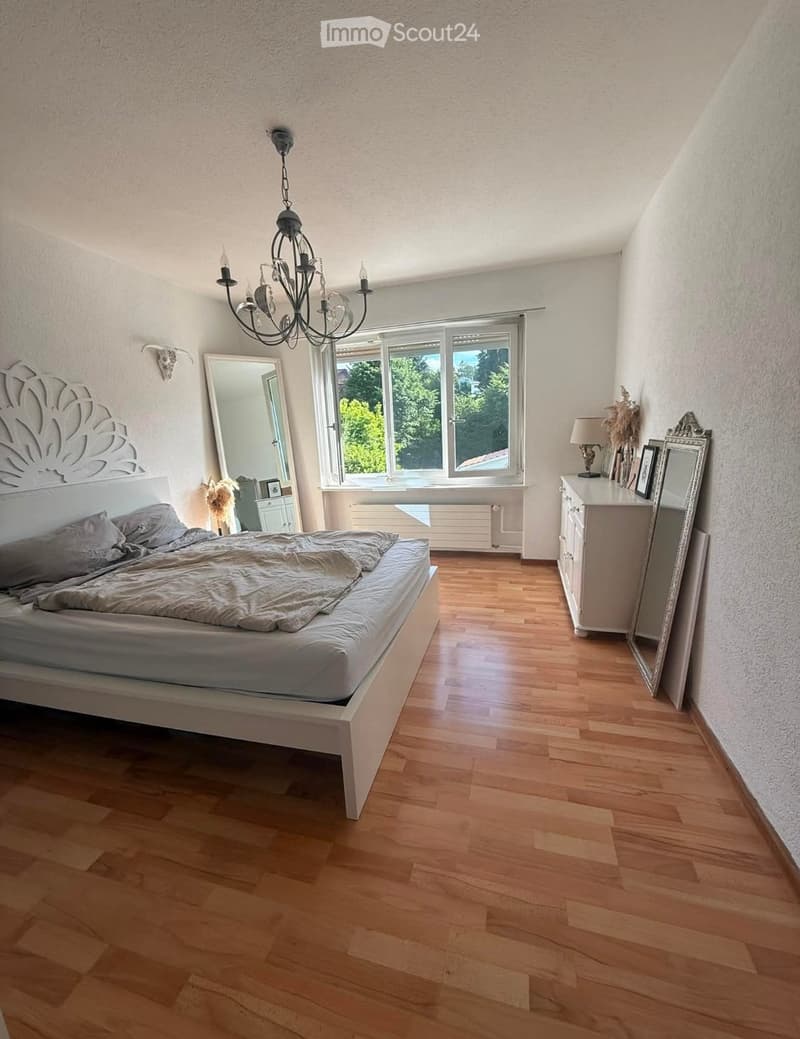 Bright bedroom with wooden flooring, large window, bed with grey bedding, chandelier, nightstand with lamp, and decorative mirror.