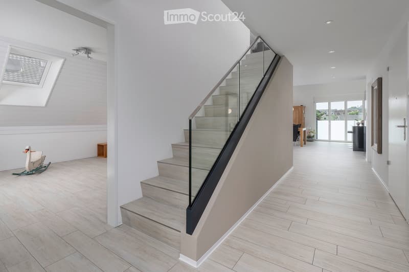Modern hallway with light wooden flooring, white walls, and a glass-enclosed staircase leading upwards. Features include recessed ceiling lights, a skylight window, and a rocking chair in the corner.