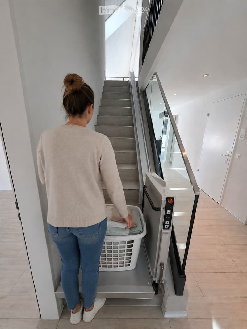A modern hallway with a staircase, glass railing, and a laundry lift. A woman is placing laundry into the lift.