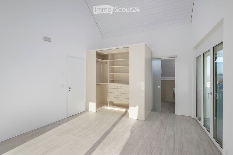 Bright, minimalist room with light wooden flooring, white walls, and a large built-in closet with open shelving and drawers. The room features a sliding glass door leading to another space and a white door on the left side.