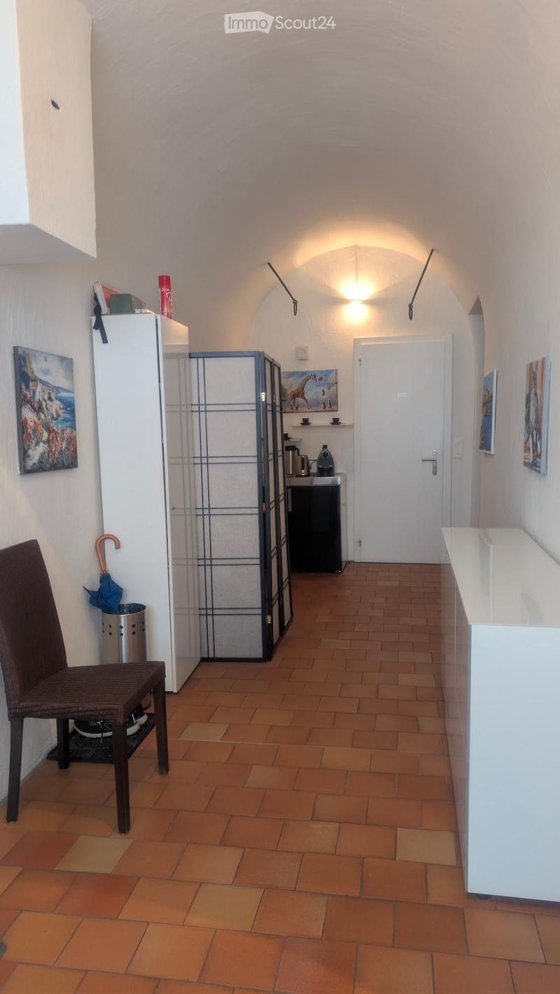 A narrow hallway with terracotta tiled flooring, white walls, and a small seating area with a wicker chair and umbrella stand. A small kitchen area is visible with a refrigerator, coffee machine, and countertop.
