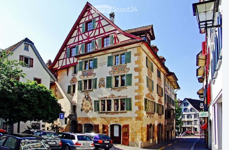 A historic building with a distinctive red and white timber-framed roof, ornate facades, and multiple windows with shutters. The building is situated on a street with parked cars and other buildings in the background.