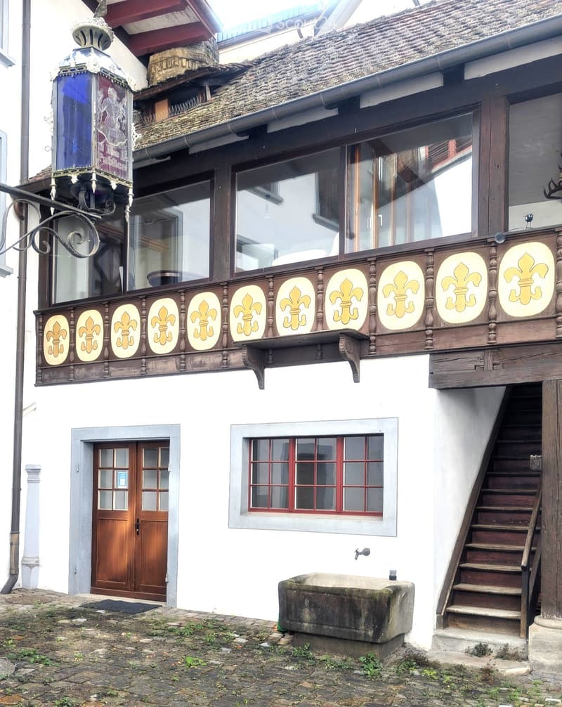 Two-story building with white walls, wooden balcony with fleur-de-lis decorations, wooden door, and red-framed window.