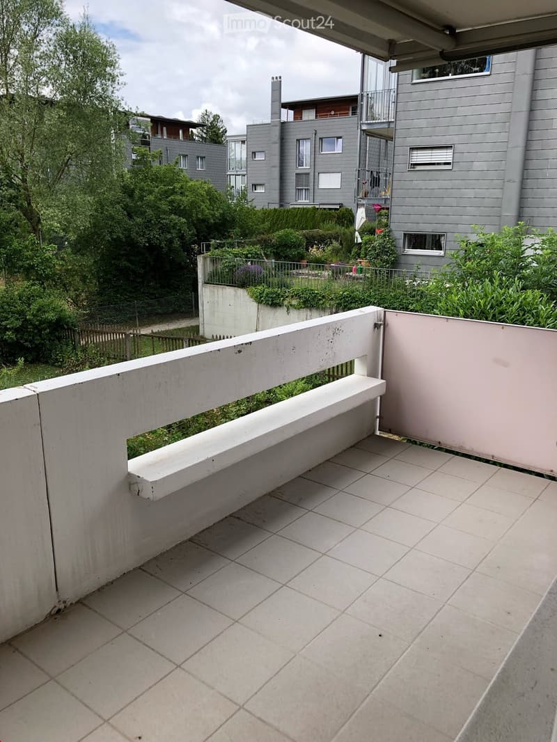 Balcony with tiled floor, white railing, overlooking a landscaped garden area with trees and shrubs.