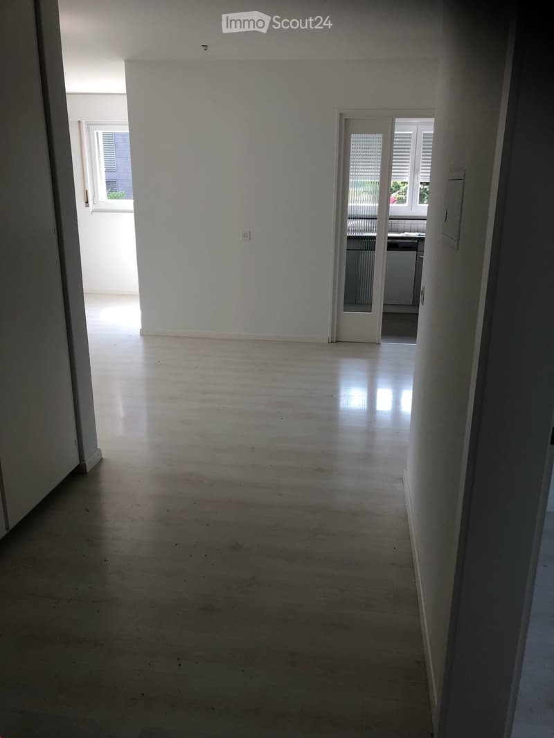 Empty hallway with light-colored walls and a polished concrete floor, leading to an adjacent room with white cabinets and a window.