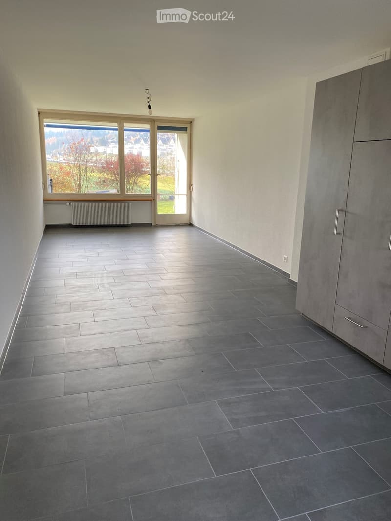 Empty room with grey tiled floor, white walls, large window with view, radiator, and built-in cabinet.