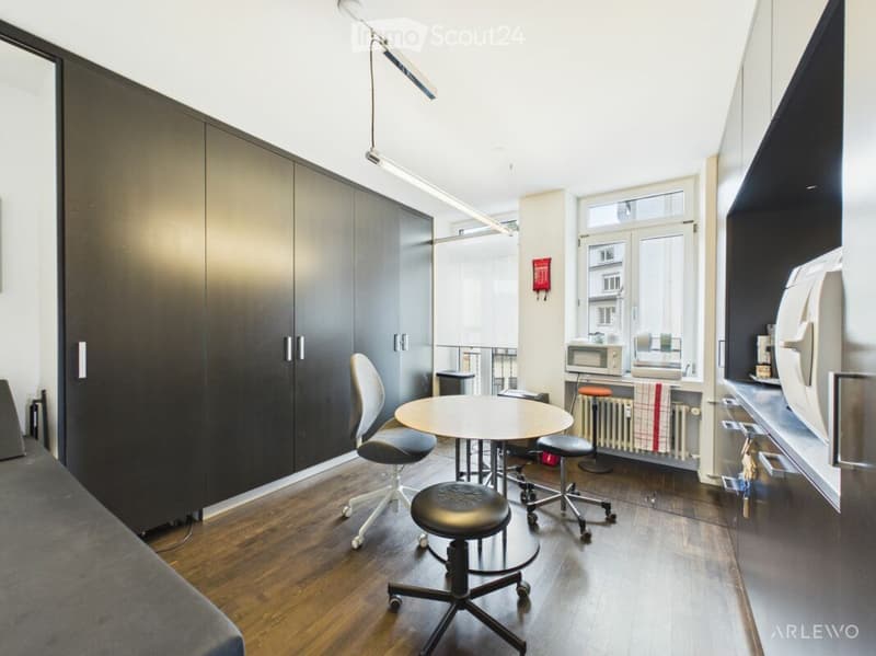 Modern, compact room with wooden flooring, large window, and a mix of furniture including a table, chairs, and a microwave.