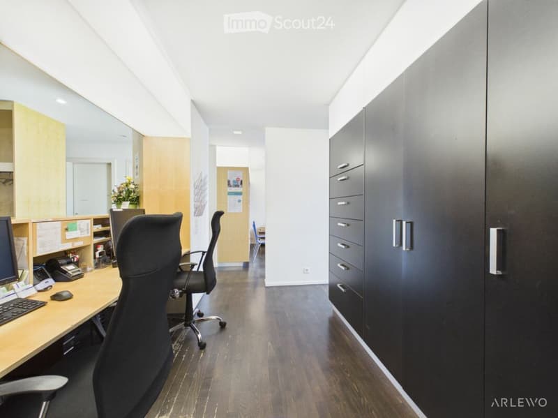 Modern office space with wooden flooring, a large desk with a computer setup, ergonomic chairs, and built-in storage cabinets.