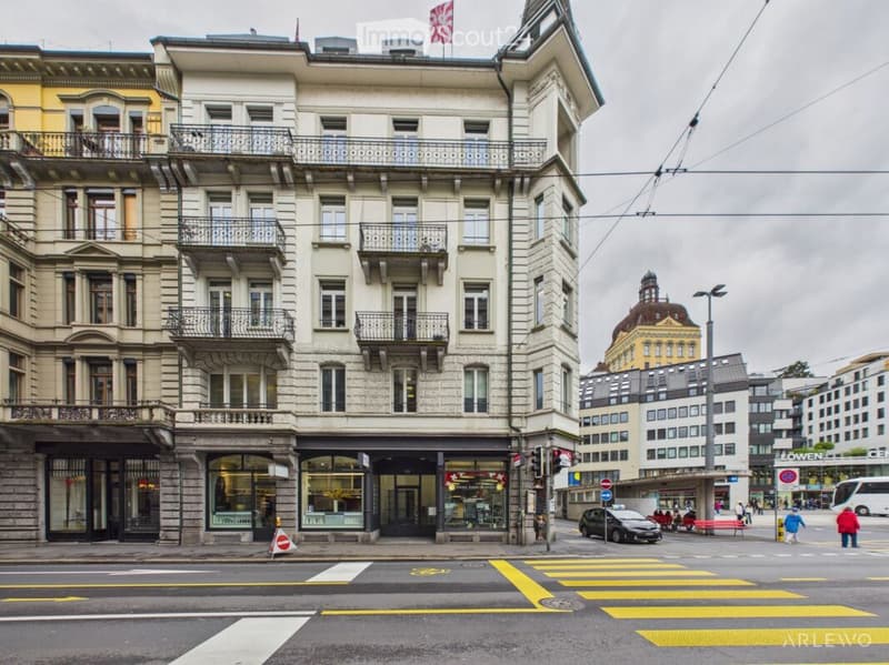 Multi-story building with balconies, large windows, and a storefront at the ground level. The building is situated on a street corner with a pedestrian crossing in the foreground.