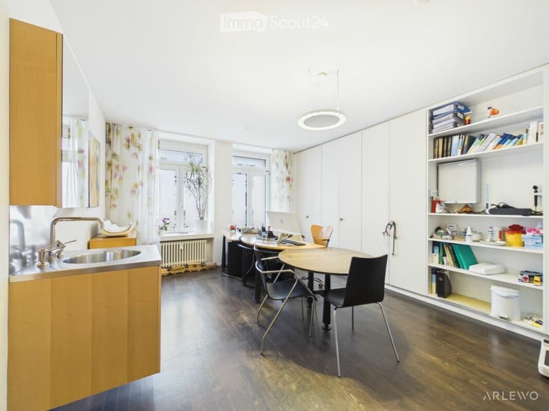 A modern, open-plan kitchen and dining area with wooden cabinets, a round dining table, chairs, a laptop, and a bookshelf filled with books and decorative items.