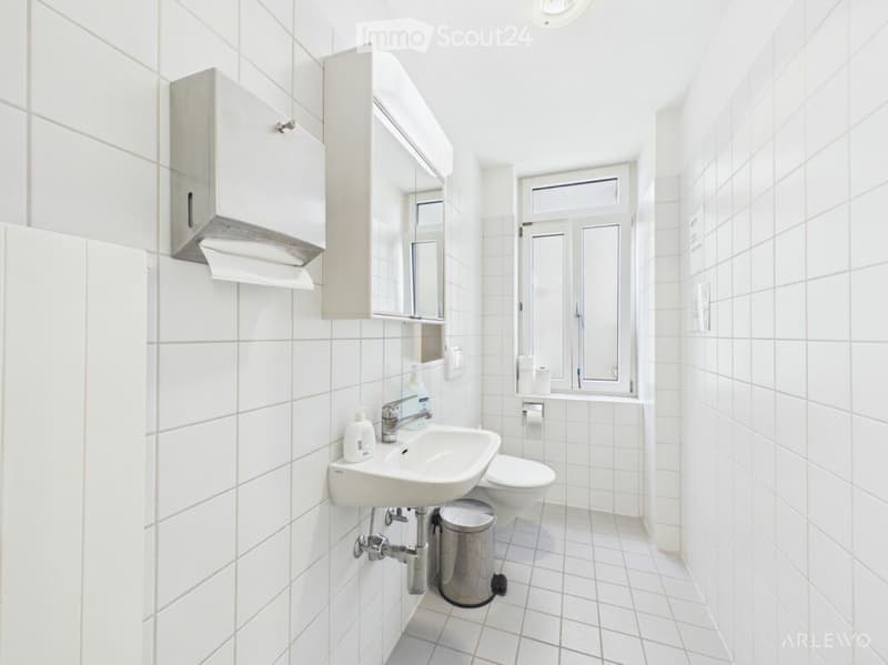 Modern bathroom with white tiles, a sink, toilet, mirror, and a small cabinet. A window provides natural light.
