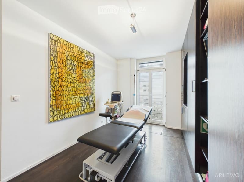 A modern, well-lit treatment room with a black treatment table, a small stool, a cabinet, a screen, and an art piece on the wall. The room features a large window with blinds and a sleek, contemporary design.