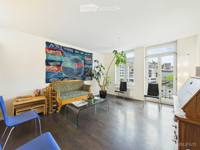 Bright living room with wooden flooring, large windows, a colorful abstract painting, a sofa, coffee table, chairs, and a potted plant.
