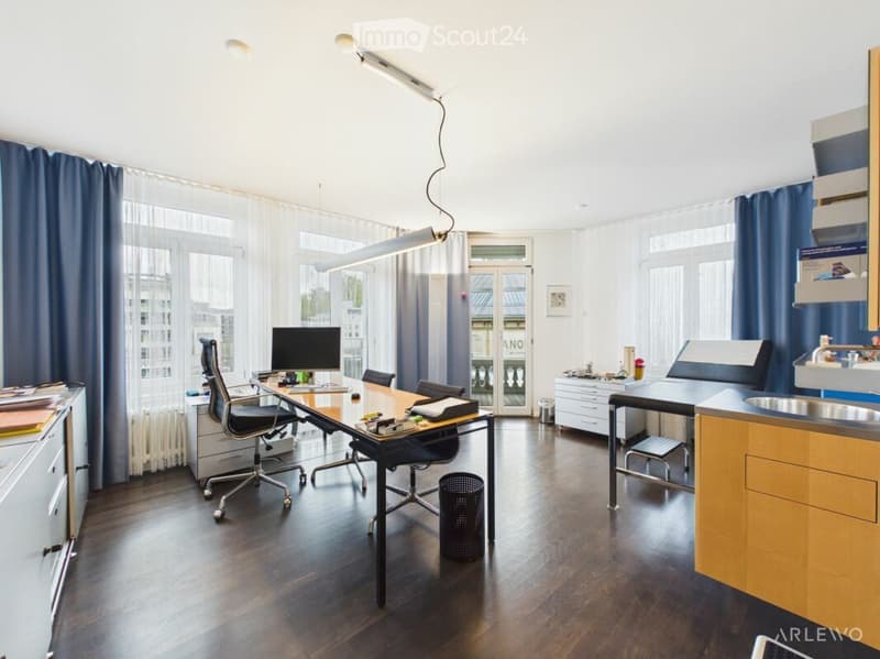 Modern office space with a large wooden desk, ergonomic chair, computer, cabinets, sink, and a small balcony area visible through large windows.