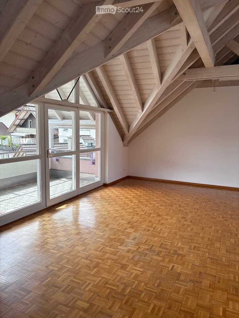 Empty attic room with wooden herringbone flooring, exposed wooden beams, and large windows offering a view of the neighborhood.