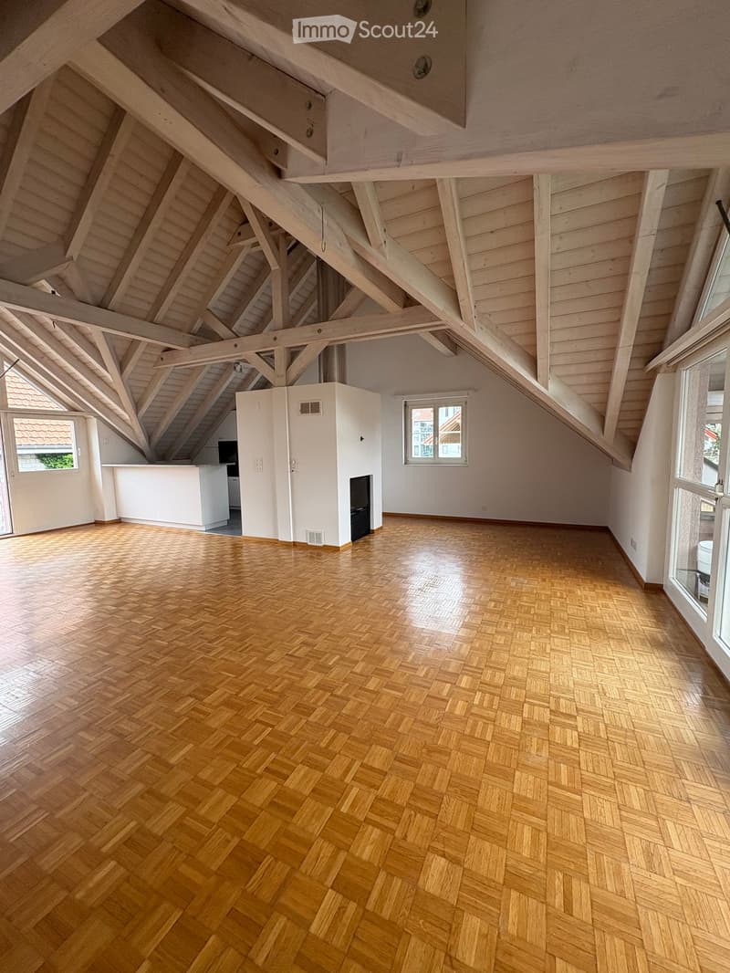 Bright, spacious attic room with wooden herringbone flooring, exposed wooden beams, and large windows offering natural light. The room features a small kitchen area with white cabinets and a black stove, and a fireplace is visible in the center.