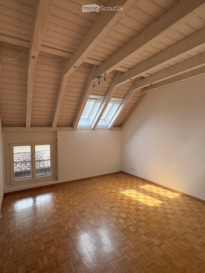 Empty attic room with wooden herringbone flooring, exposed wooden beams, skylights, and a window with a metal railing view of a tiled roof.