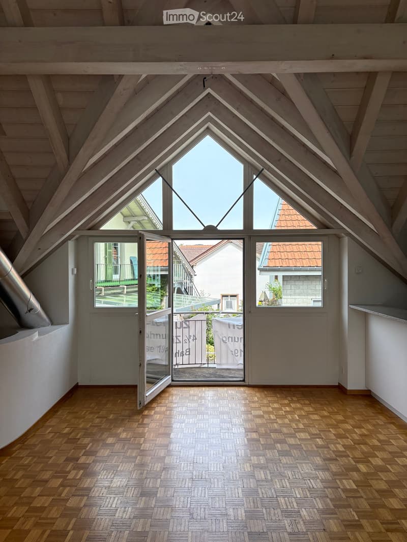 Bright, open attic space with wooden herringbone flooring, exposed wooden beams, large skylights, and a set of glass doors leading to a balcony.