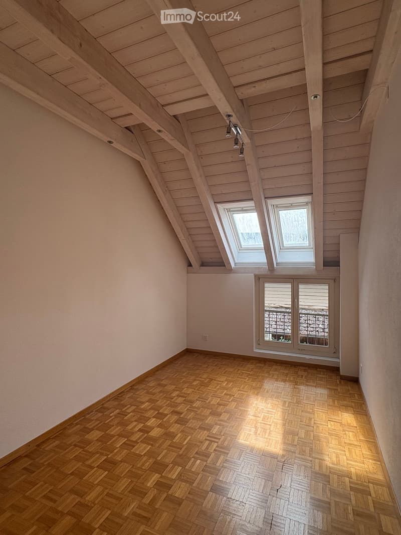 Empty attic room with wooden herringbone flooring, exposed wooden beams, skylights, and a window with a view of a stone wall and railing.