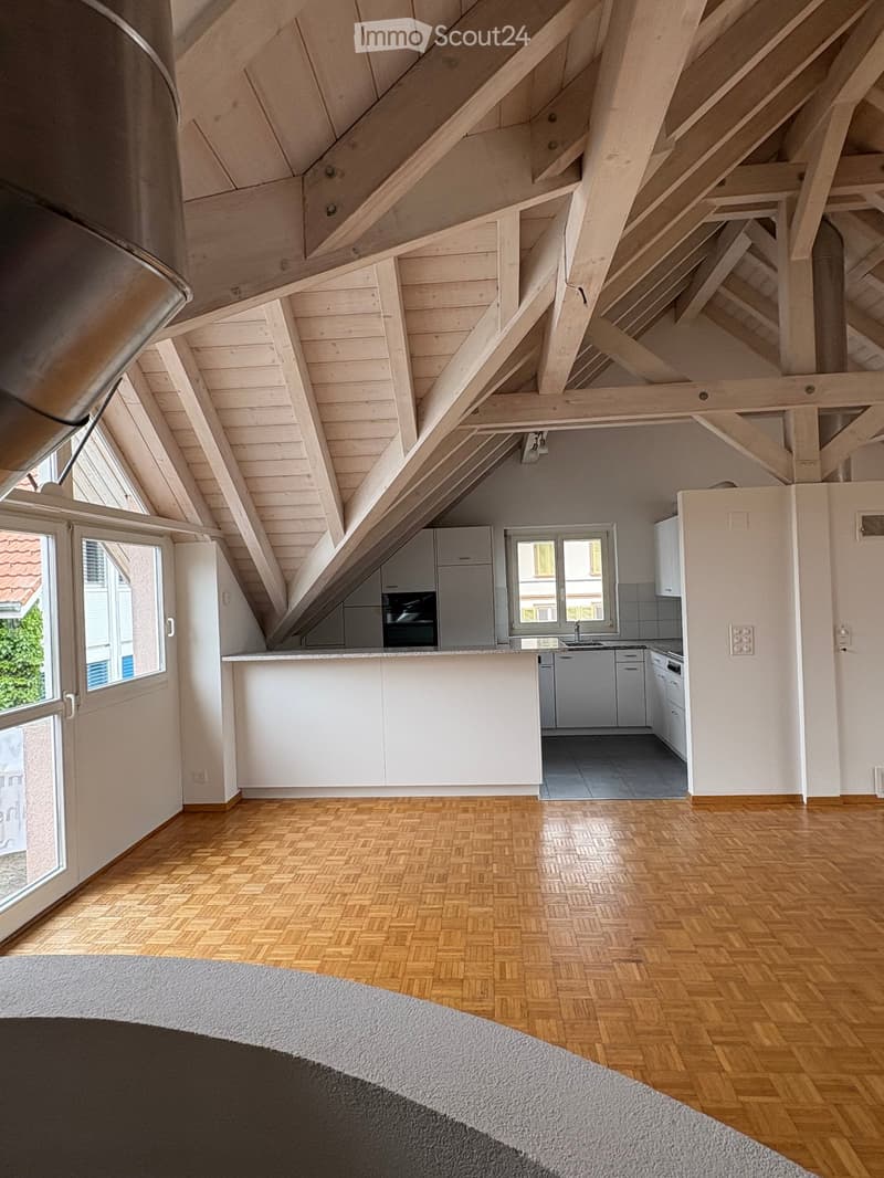 Bright, airy attic space with exposed wooden beams and slanted ceilings. Features a modern kitchen area with white cabinets, a central island, and stainless steel appliances, including an oven, refrigerator, and dishwasher. The room has large windows, woo