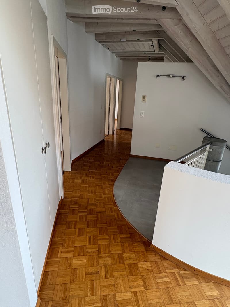 Spacious hallway with wooden herringbone flooring, white walls, and a curved staircase leading upwards. Features include a metal railing, a small trash bin, and several electrical outlets.