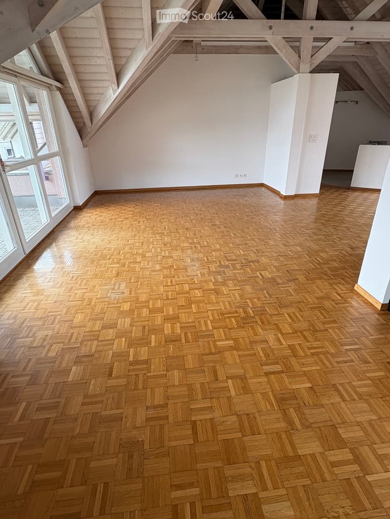 Empty room with wooden herringbone flooring, white walls, and large windows with wooden frames. Exposed wooden beams on the ceiling.