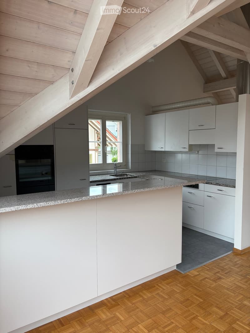 Modern kitchen with white cabinets, granite countertop, sink, faucet, and built-in oven. Wooden herringbone floor and slanted wooden ceiling.