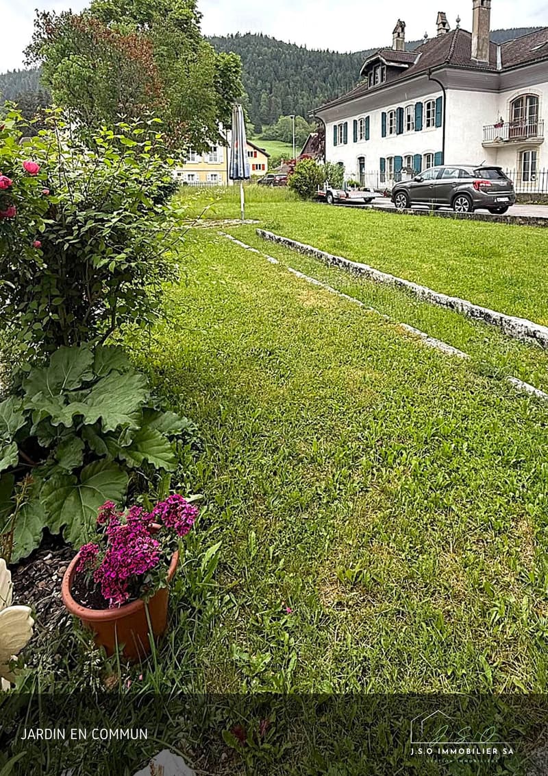 Well-maintained garden with green grass, potted flowers, and a pathway leading to a large house with multiple windows and a balcony. The house is surrounded by trees and other residential buildings in the background.