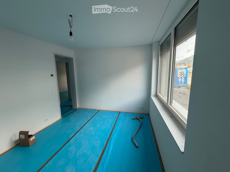 Empty room with light blue protective flooring, white walls, large window, and a small open doorway leading to another space.