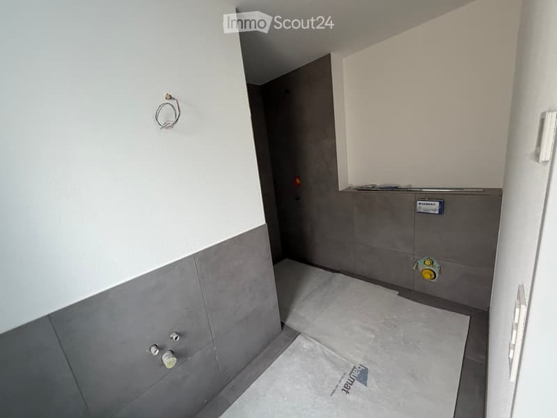 Empty room with grey tiled walls and floor, visible electrical outlets and a water valve.