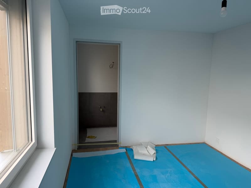Empty room with light blue walls, a large window on the left, and a blue protective floor covering. A small closet with shelves is visible in the background.