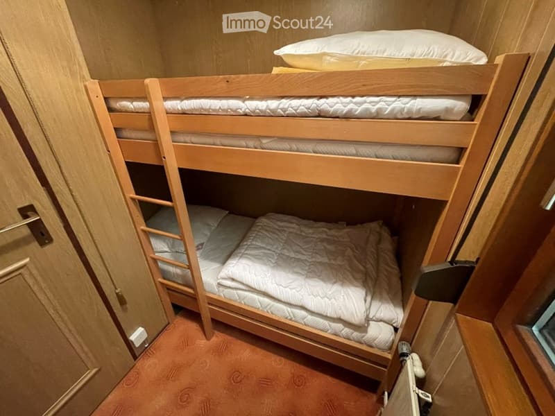 Compact bedroom with a wooden bunk bed, neatly made with white bedding and pillows, set against light-colored walls and a warm-toned carpet.