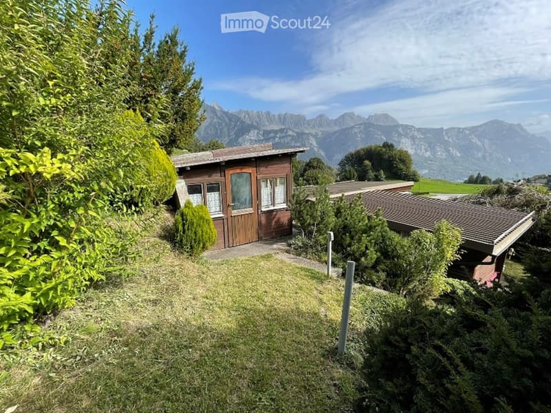 A small wooden cabin surrounded by lush greenery and trees, with a scenic mountain view in the background.