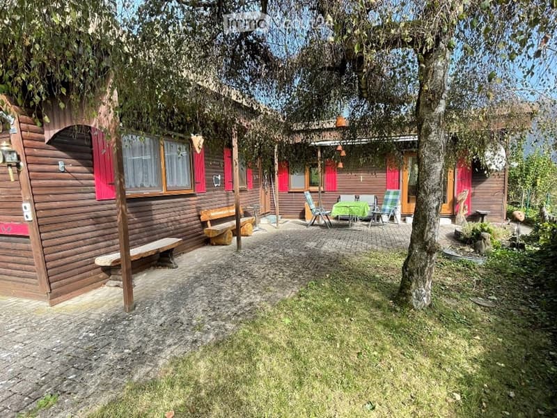 Wooden house with red shutters, outdoor seating area with benches and chairs, green tablecloth, and a tree providing shade.