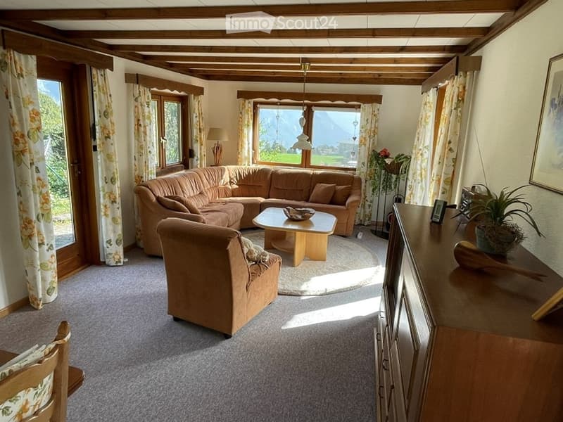 Bright living room with large windows, wooden beams on the ceiling, a sectional sofa, coffee table, armchair, and a side table with a lamp and plant.