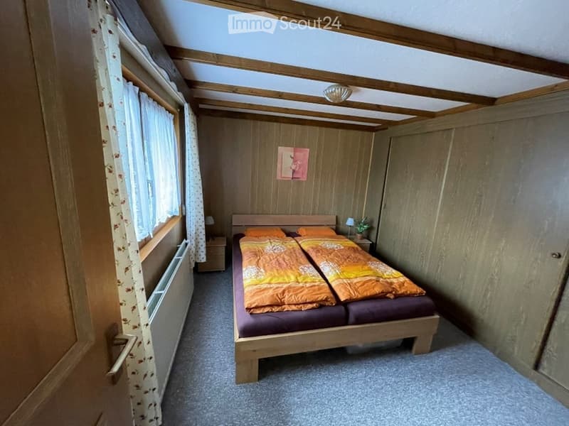 Cozy bedroom with wooden walls and ceiling beams, featuring a double bed with orange and purple bedding, nightstands with lamps, and a small plant on one nightstand.