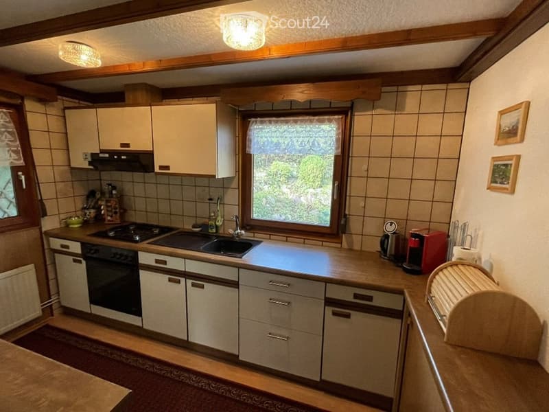 A cozy kitchen with white cabinets, a stove, sink, and various appliances including a coffee maker and a dish rack. The space features tiled walls and a window with a view of greenery outside.