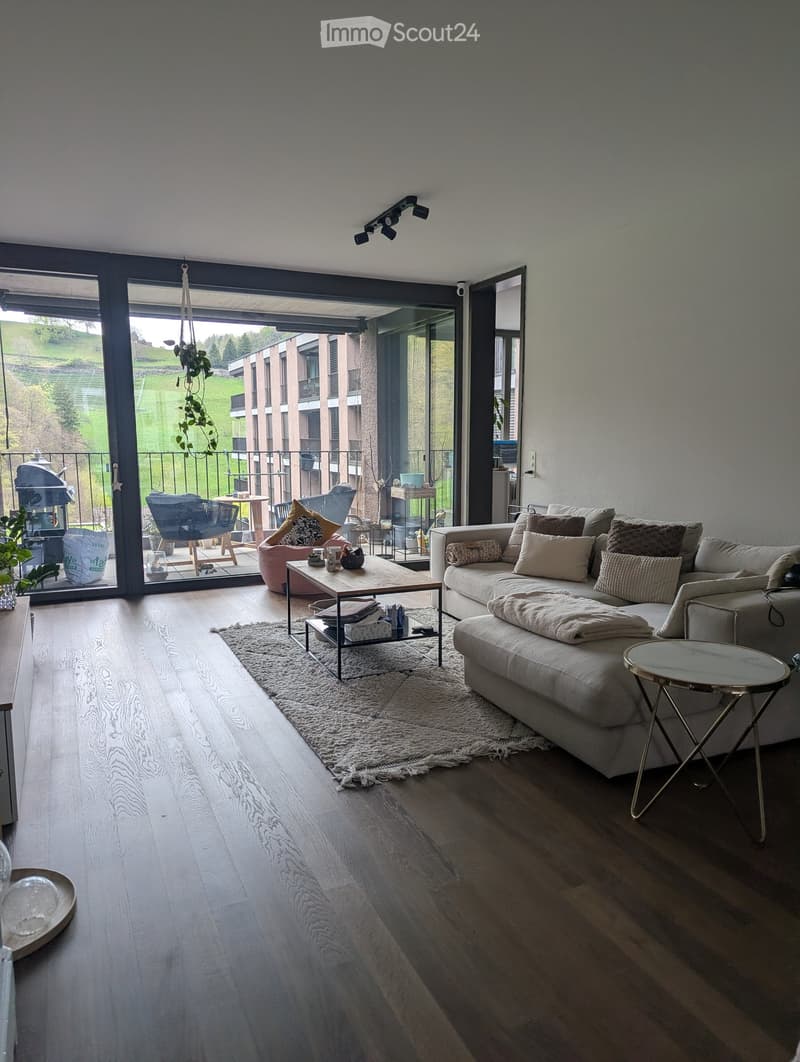 Modern living room with large glass doors leading to a balcony, featuring a light grey sofa, coffee table, side table, and a small round table. Wooden flooring and a white shag rug are present.
