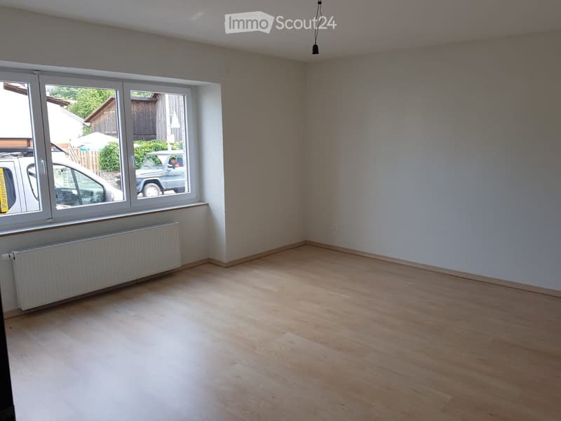 Empty room with wooden flooring, white walls, large window with radiator below, and visible outdoor area with parked cars.