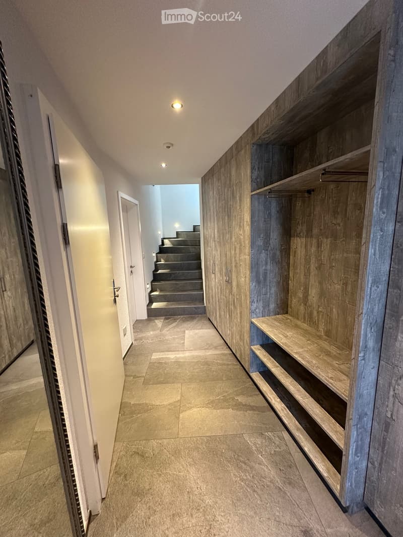 Modern hallway with tiled flooring, built-in wooden shelving unit, recessed lighting, and a staircase leading upwards.
