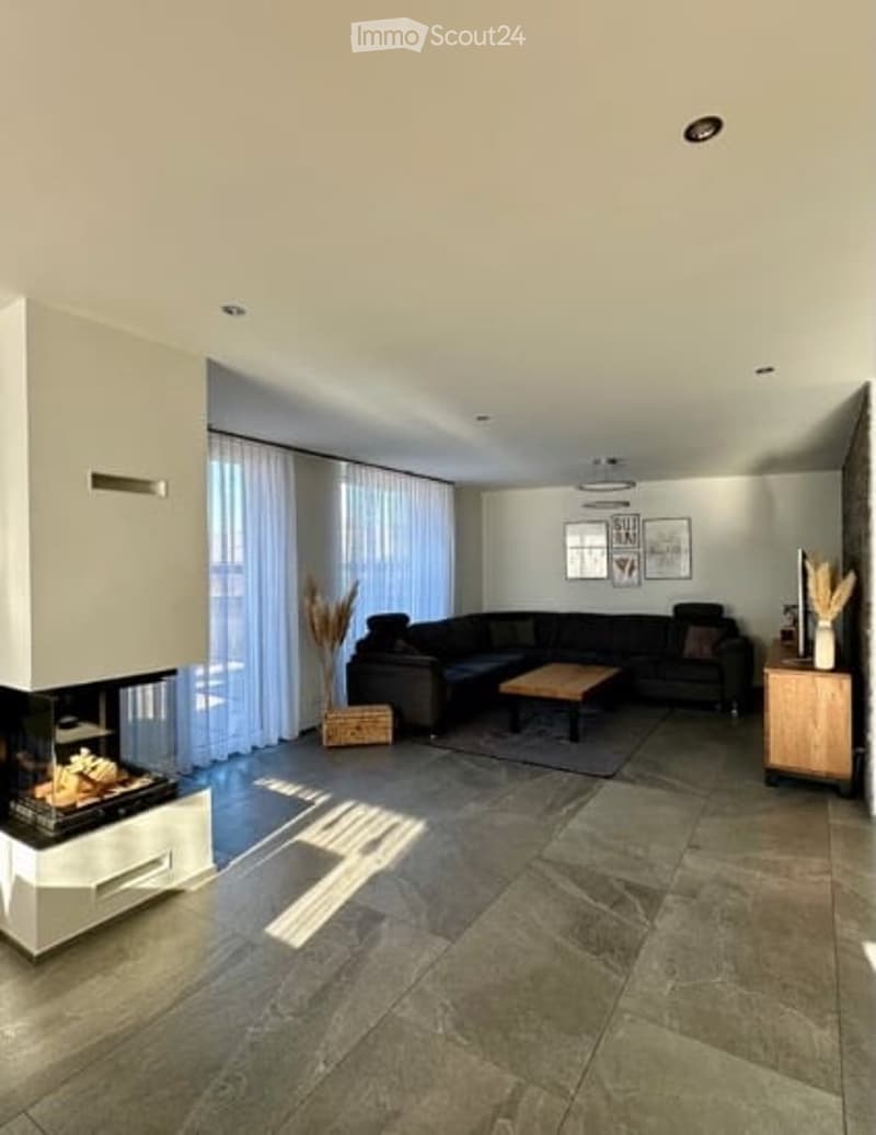 Modern living room with a dark sectional sofa, coffee table, fireplace, side table, and decorative vases. Large windows with white curtains allow natural light to fill the space.
