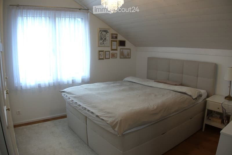 Cozy bedroom with a large bed, tufted headboard, nightstand, lamp, and several picture frames on the wall. The room features wooden paneling and a slanted ceiling with a chandelier.