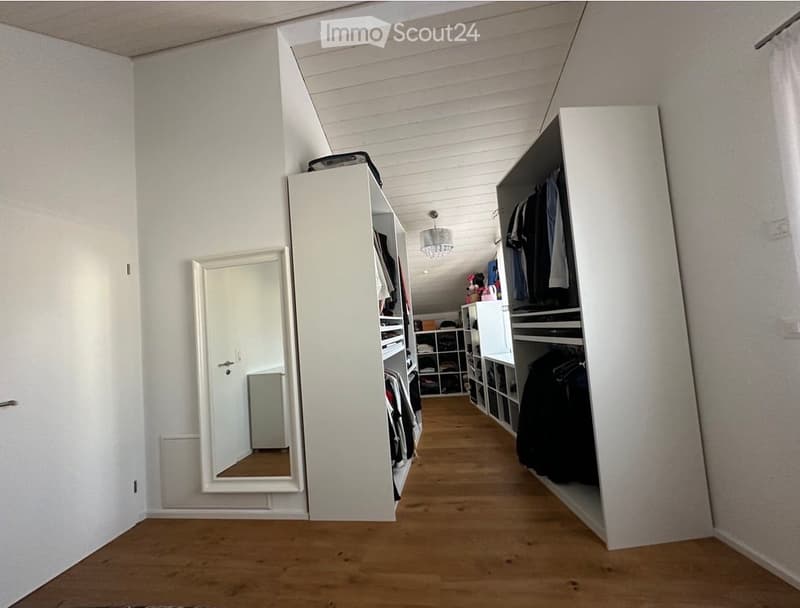 Spacious walk-in closet with built-in wardrobes, shelves, and a full-length mirror. The room features light wooden flooring and white walls.