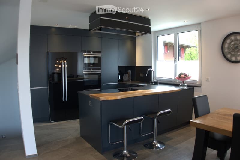Modern kitchen with dark grey cabinets, stainless steel appliances, wooden countertop, bar stools, and a small dining area with a table and chairs.
